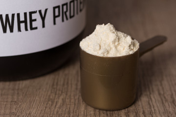 Whey Protein. Golden scoop with vanilla flavour powder and black jar of supplement. Wooden table. Close up, texture.