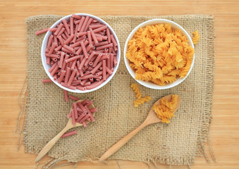 Spiral Macaroni and red pasta in white bowl on sack against wood background.