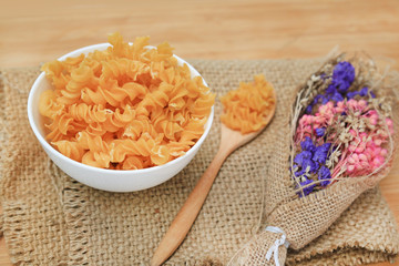 Spiral Macaroni in white bowl on sack decoration with dry flower bush against wood background.