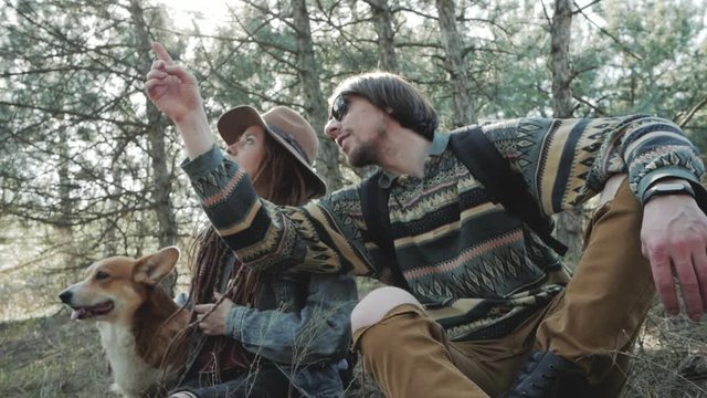 Young couple with backpacks in spring forest, hikers with corgi dog relaxing in sunny forest 