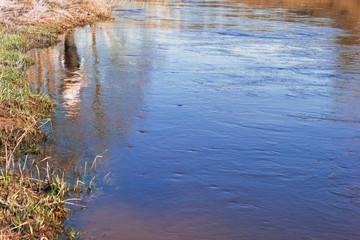 Calm flow of a flat river with the reflection of a fisherman standing on the shore. A small ripple on the water. Sunny spring morning.