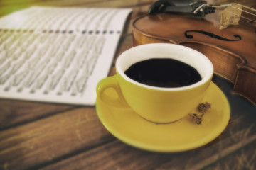 In selective focus of yellow ceramic coffee cup put beside violin ,in wooden timber board,blurry light design background.