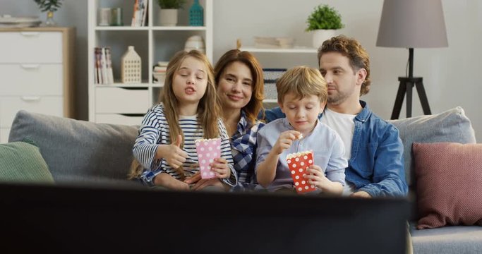 Cheerful caucasian family eating popcorn while sitting on the couch in the living room and watching TV. Portrait shot. Inside