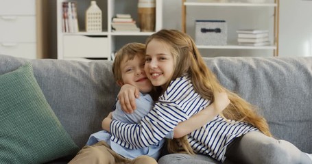 Portrait shot of the cute small sister and brother sitting on the couch in the living room, hugging each other and posing in front of the camera. Indoors