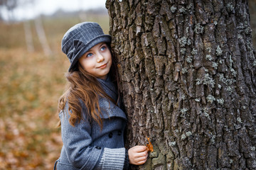 A cute little girl in grey coat and gray hat near the thick old oak in the oak wood at the sunset