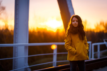 Obraz premium A little girl in a yellow coat walking on the railway iron bridge at the sunset in a cold evening