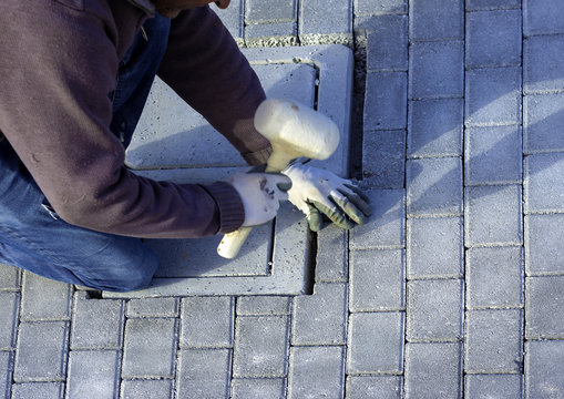 Construction worker laying interlocking paving concrete onto sheet nonwoven bedding sand and fitting them into place.