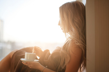 Young beautiful Girl drinking coffee sitting near the window in morning. Blond sexy naked female...