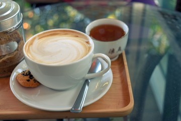 Latte coffee in a white cup with cookie and tea on a transparent glass table
