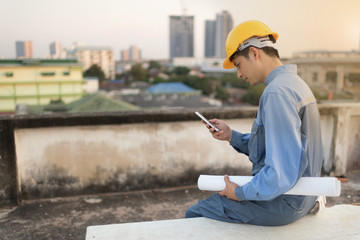 Young Asian technician man using smartphone with safety helmat and holding plans.