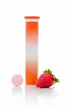 Tube of sparkling tablets with one piece next to tube. Pipe with fizzy tablets with vitamin c. Pastils with straberry flavour with fresh fruit next to. Healing tabletes in tube and one piece outside