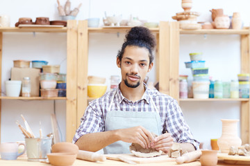 Fototapeta na wymiar Portrait of young confident mixed race man modeling clay pot in workshop and looking at camera