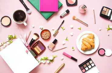 Fototapeta na wymiar Various cosmetic products for makeup and white spring flowers in a white gift bag, notepad, cup of coffee, croissant on a pink pastel background top view with copy space