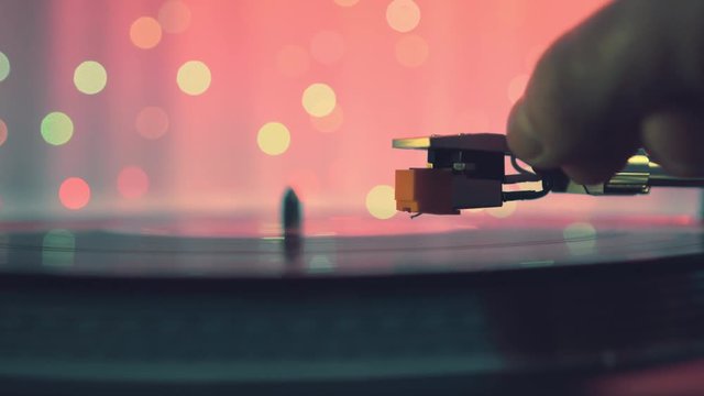The moment of a rotating vinyl plate turntable on a pink background and bright bokeh lights. Sound technology for DJ to mix and play music. The man's hand puts and removes needle from the vinyl plate 