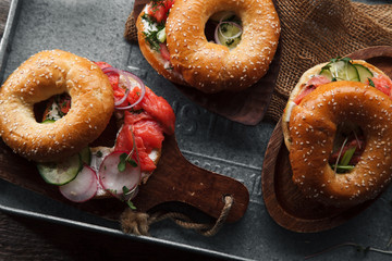 Bagels with salmon fish, cream cheese, cucumber and fresh radish slices on metallic tray on rustic gray wooden background