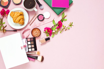 Various cosmetic products for makeup and pink spring flowers in a white gift bag, notepad, cup of coffee, croissant on a pink pastel background top view with copy space