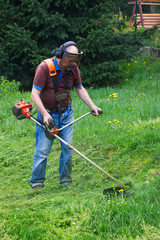 Senior gardener mowing grass by brushcutter in garden 