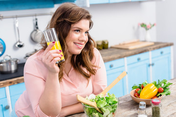 side view of overweight smiling woman with glass of juice in kitchen at home