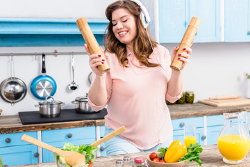 cheerful overweight woman in headphones with wooden salt and pepper grinders in hands in kitchen