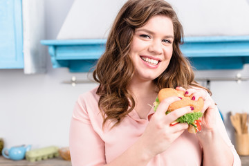 portrait of overweight smiling woman with burger in hands in kitchen at home