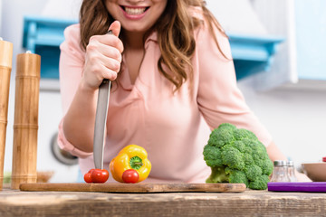 partial view of smiling woman with knife at table with fresh vegetables in kitchen at home