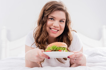 portrait of overweight smiling woman in pajama with burger on bed at home