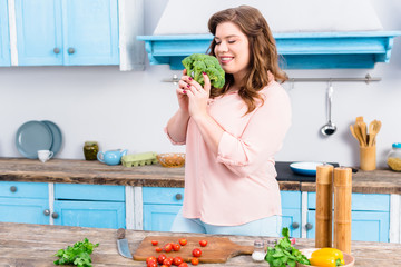 portrait of overweight smiling woman with fresh broccoli in hands in kitchen at home