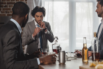 cropped shot of multiethnic men drinking alcohol and looking at friend smoking cigar