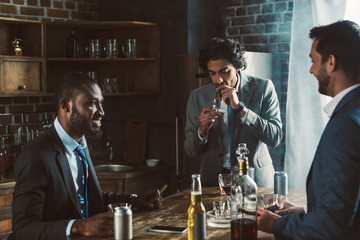 smiling young male friends in formal wear smoking cigars and drinking alcoholic beverages while...