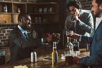cropped shot of smiling multiethnic male friends in suits drinking alcoholic beverages and smoking cigars together