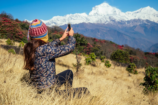 a young tourist woman with a hiking backpack and a knitted hat taking pictures of the landscapes and making selfi in the Himalaya mountains. trekking concept in the mountains