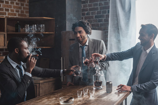cheerful male friends in formal wear smoking cigars and clinking glasses of whiskey