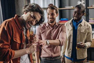 smiling multiethnic men looking at friend opening beer bottle with teeth