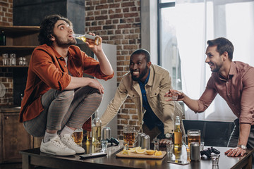 smiling multiethnic men looking at friend drinking beer while crouching on table