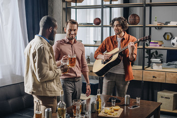 happy multiethnic male friends drinking beer and playing guitar together