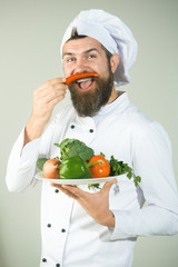 Male chef in white uniform holds pepper beside face. Bearded chef isolated on grey background. Cooking and vegetarian diet concept. Bearded chef holds plate with vegetable.