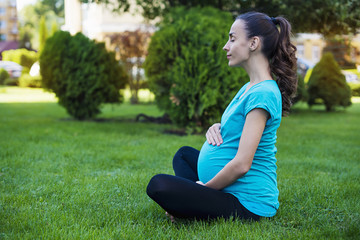 Fototapeta premium Yoga for pregnant women. Beautiful smiling young pregnant woman sitting on grass in a lotus pose and doing special breathing exercises