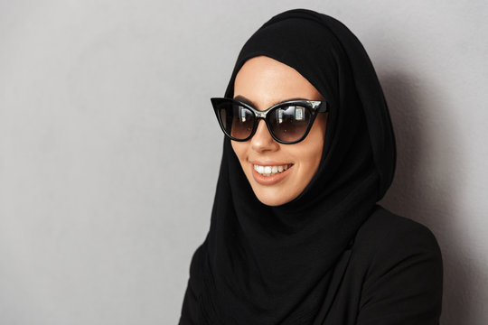 Portrait closeup of muslim fashion woman 20s in religious headscarf and sunglasses smiling and looking aside, isolated over gray background