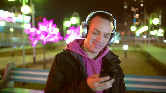 Smiling and laughing handsome young man listening music from his smartphone in wireless black headphones, dancing on street of old town center in europe at twilight or evening time. 4 k
