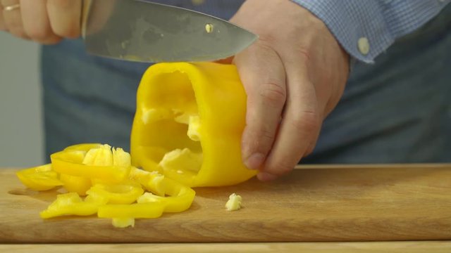 male hands holding sharp knife and chopping fresh yellow pepper on wooden board