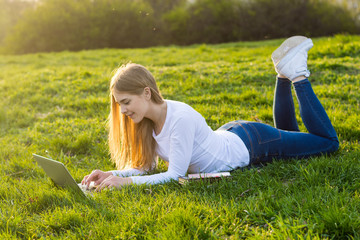 Young beautiful woman with long blond hair lies with a laptop on the grass in spring in sunset.