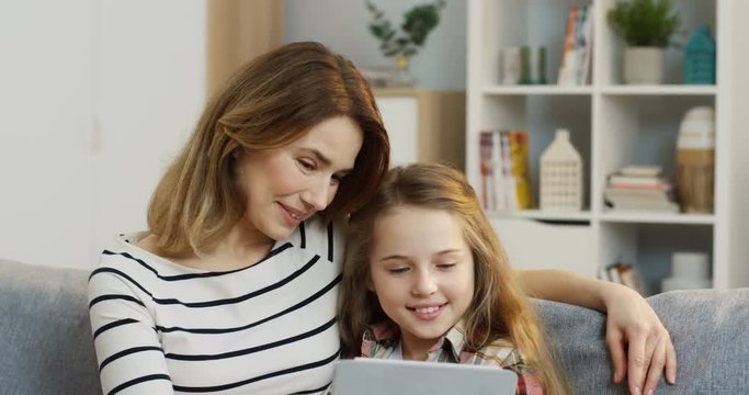 Close up of the beautiful mother sitting on the sofa and hugging her pretty teen daughter while they watching something on the tablet device. At home. Inside