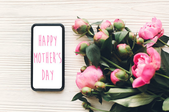 happy mother's day text on phone screen and pink peonies on rustic white wooden background in light. floral greeting card concept, flat lay. mothers day. tender spring image