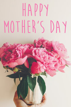happy mother's day text and hand giving pink peonies bouquet at rustic white  background in light. floral greeting card concept. mothers day. tender spring image