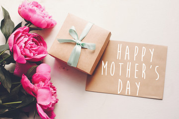 happy mother's day text on craft card and pink peonies bouquet with gift box on rustic white wooden...