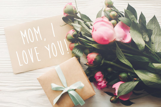 mom i love you text on craft card and pink peonies bouquet with gift box on rustic white wooden background in light. floral greeting card concept, flat lay. happy mother's day