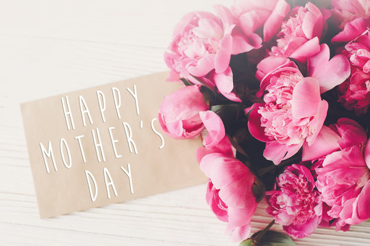 happy mother's day text on craft card and pink peonies bouquet on rustic white wooden background in light. floral greeting card concept, flat lay. mothers day. tender spring image