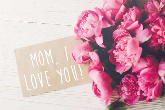 mom, i love you text on craft card and pink peonies bouquet on rustic white wooden background in light. floral greeting card concept, flat lay. happy mother's day . tender image