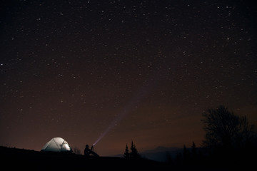 tourist with flashlight near his camp tent under a sky full of stars, night mountain loneliness