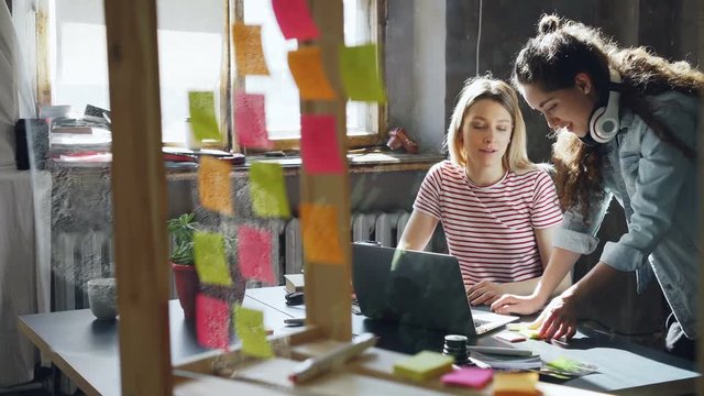 Creative team of female designers are working on new project, sharing ideas, writing information on colored stickers. Glass office wall with bright memos is in foreground.